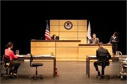 [ai] A courtroom scene with two attorneys seated at desks, a judge presiding at the front, and a witness on the stand. The American flag is displayed. The setting has a modern design with wooden panels.